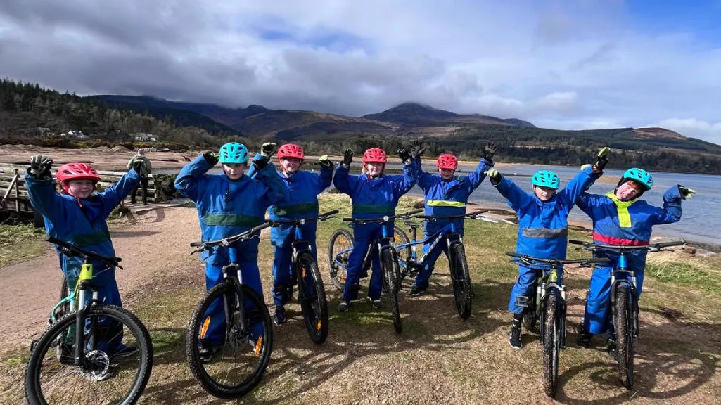 Group of children on bikes with arms raised with hills and water behind them