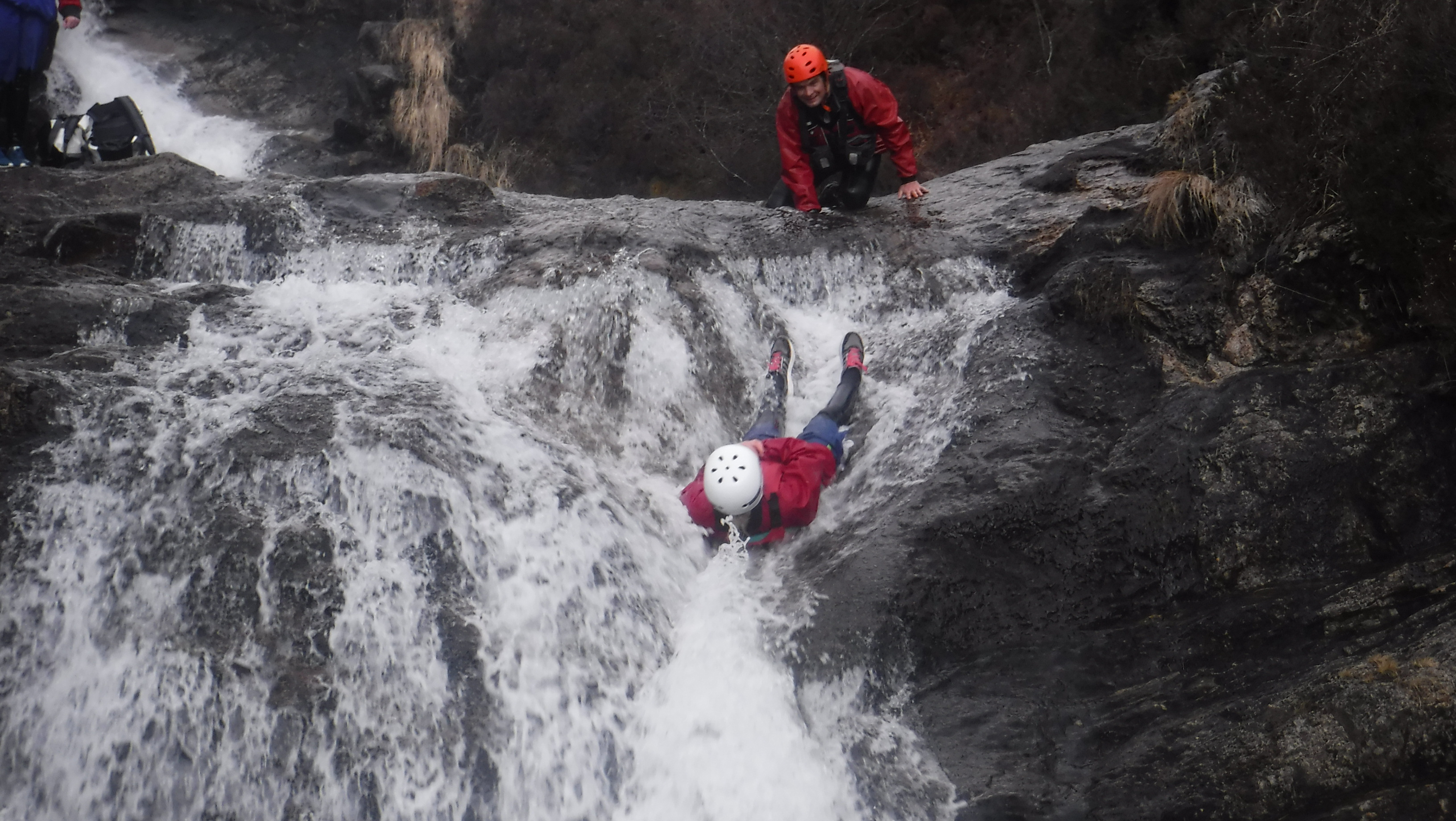 Two people in protective gear. One of whom is sliding down a waterfall.