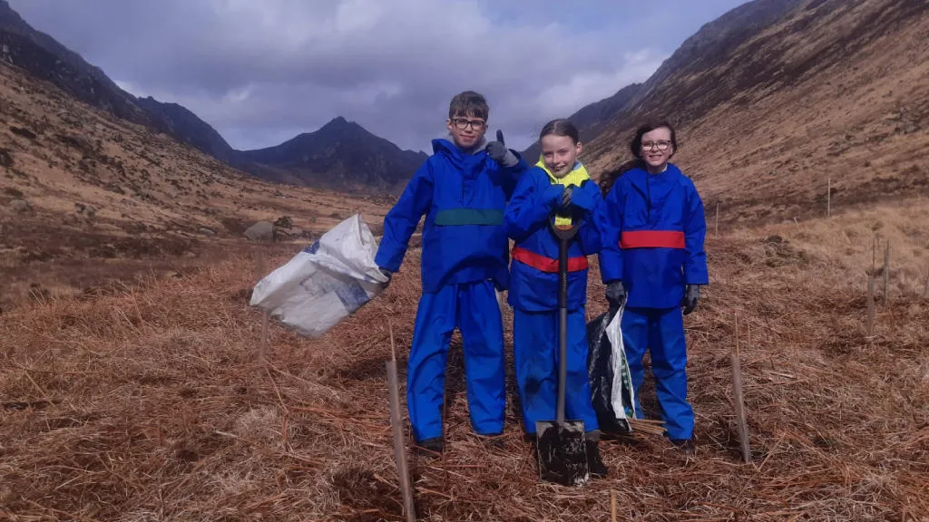 3 children standing with a shovel and plastic bag in front of hills.