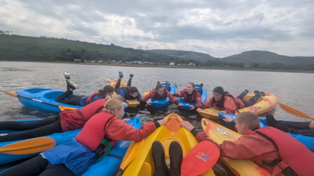 Group of teens lying on their fronts on individual kayaks while holding hands in a circle.